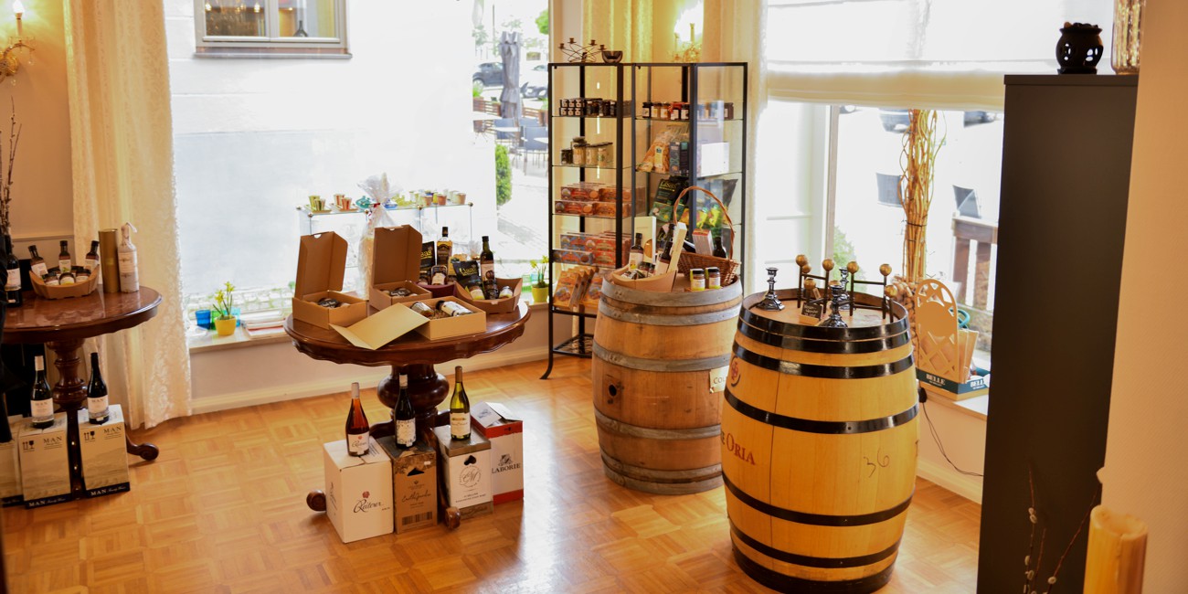 Innenaufnahme eines Feinkostladens mit großen Fenstern. In der Mitte stehen dekorativ arrangierte Holztische und Fässer mit ausgewählten Produkten. Im Hintergrund sind Regale und Pflanzen zu sehen.